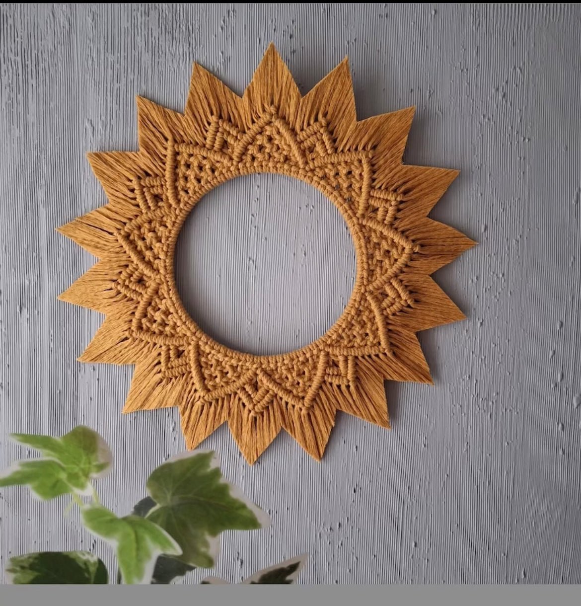 A circular, sun-shaped decorative wooden frame with intricate mandala patterns hangs on a textured, grey wall. In the bottom-left corner, there is a green plant with large leaves slightly coming into the frame, adding to the Macrame Art vibe.