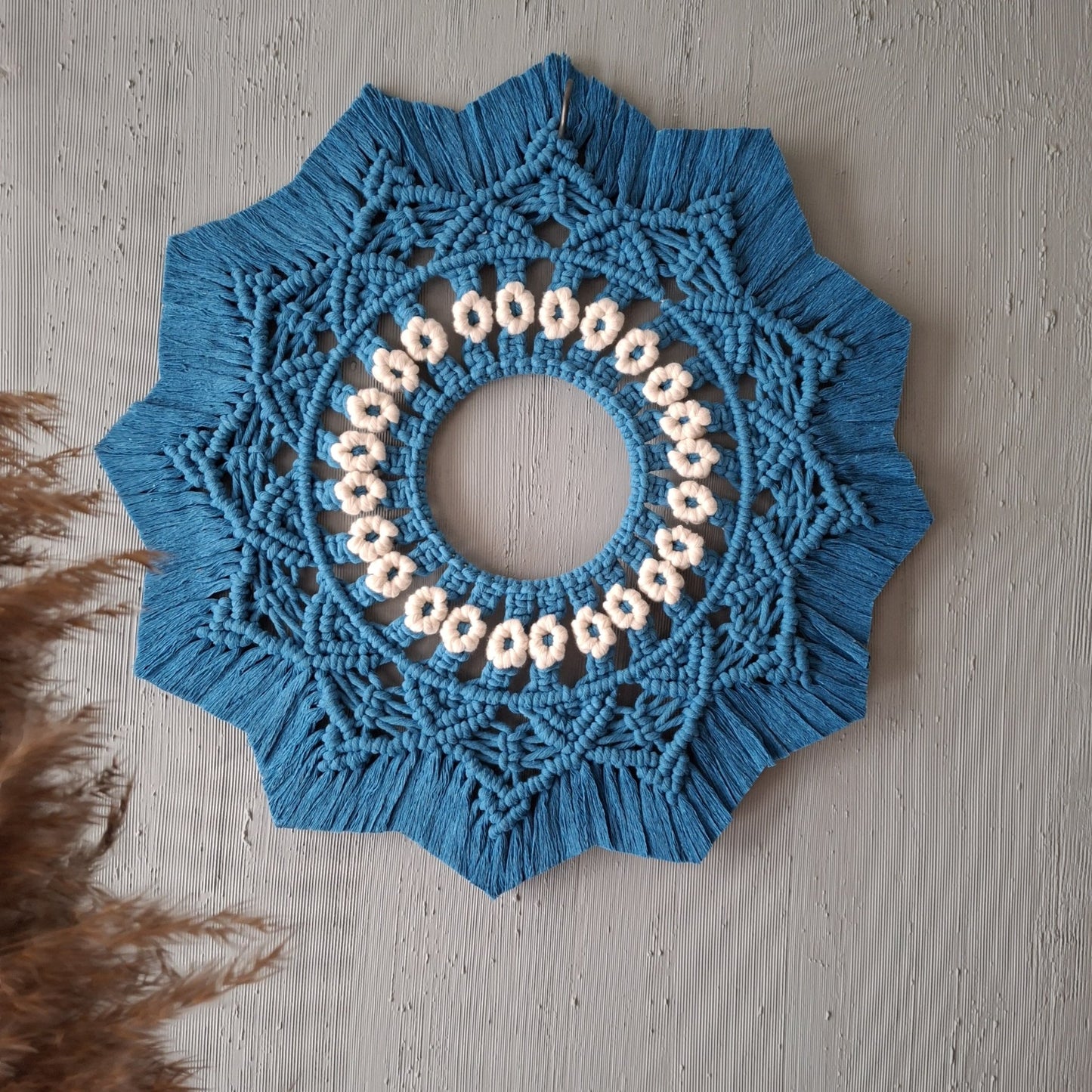 A circular, sun-shaped decorative wooden frame with intricate mandala patterns hangs on a textured, grey wall. In the bottom-left corner, there is a green plant with large leaves slightly coming into the frame, adding to the Macrame Art vibe.